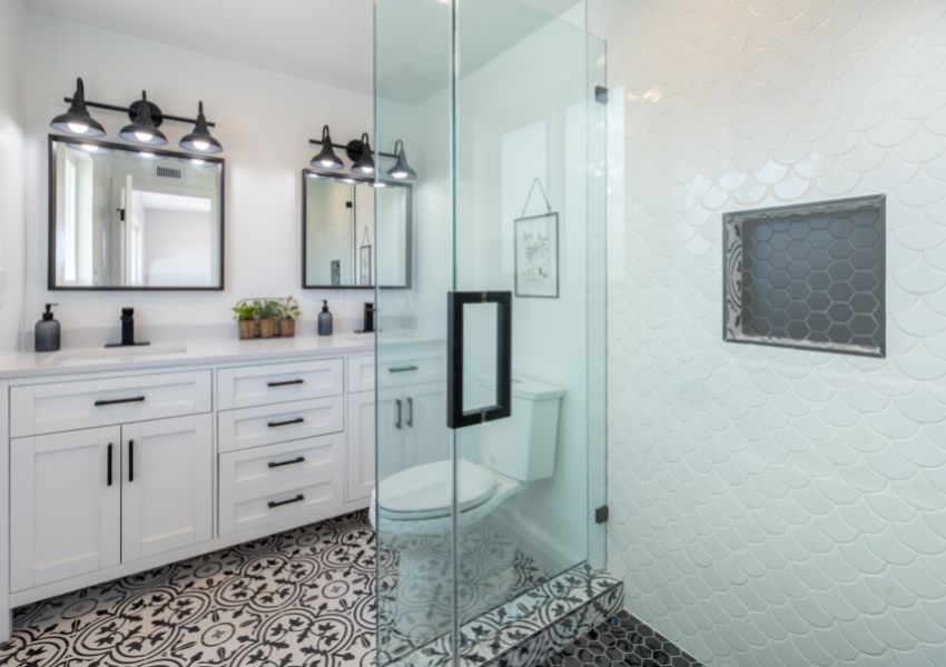 Interior of a white bathroom with black and cream tiled floors, a walk in shower with a glass door, and two sinks with square mirrors above them.